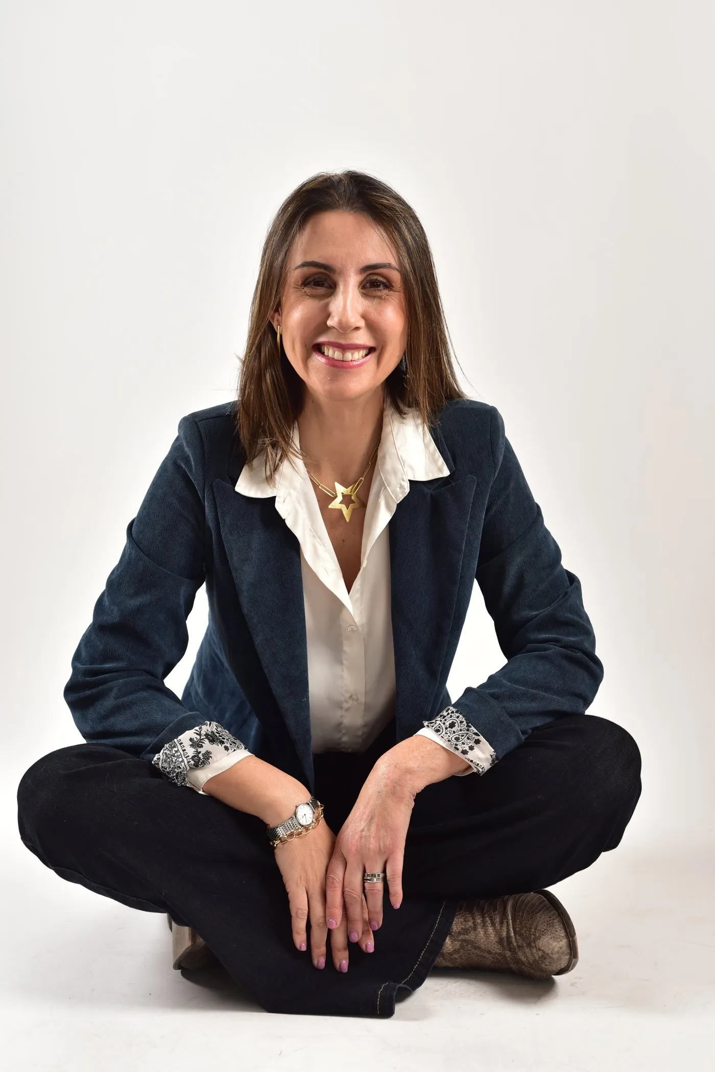 Carolina Vega sonriendo, sentada en el suelo con las piernas cruzadas, con blazer azul y blusa blanca.